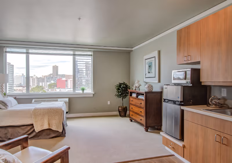 Studio apartment interior with a bed, wooden dresser, small kitchenette and a large window showing a city view.