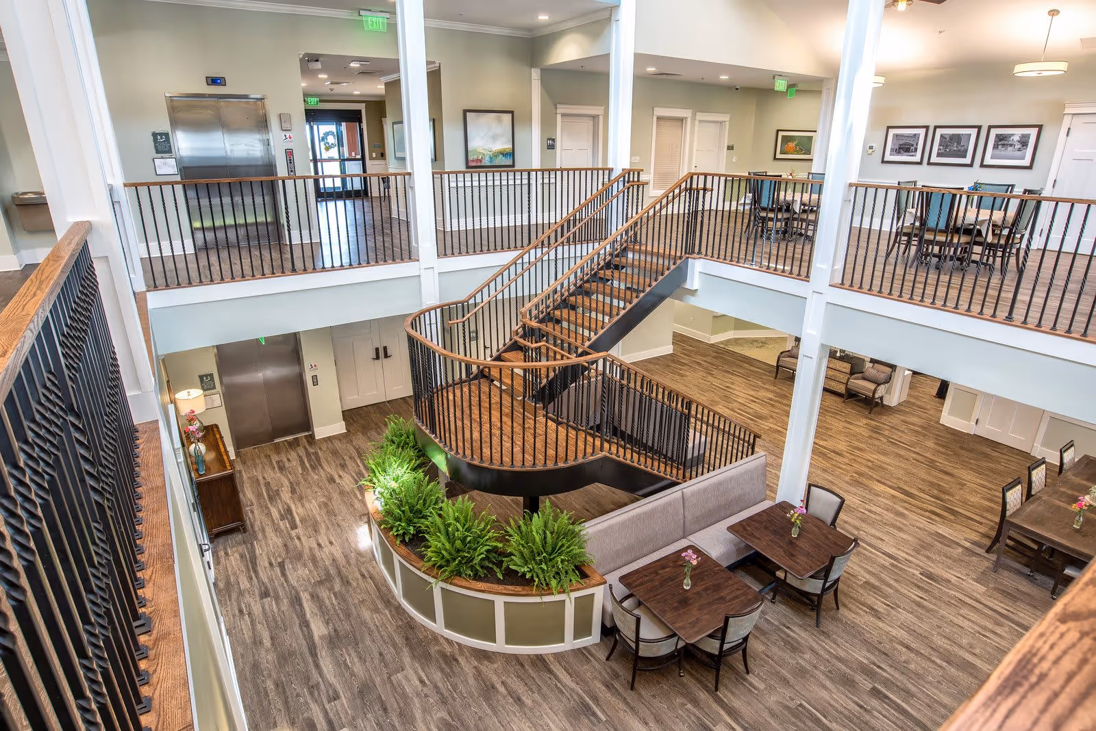 Interior view of a senior living facility showing a two-story open area with a central staircase, wooden floors, seating areas with tables and chairs, potted plants, and framed artwork on the walls. There are elevators and exit doors visible in the background.
