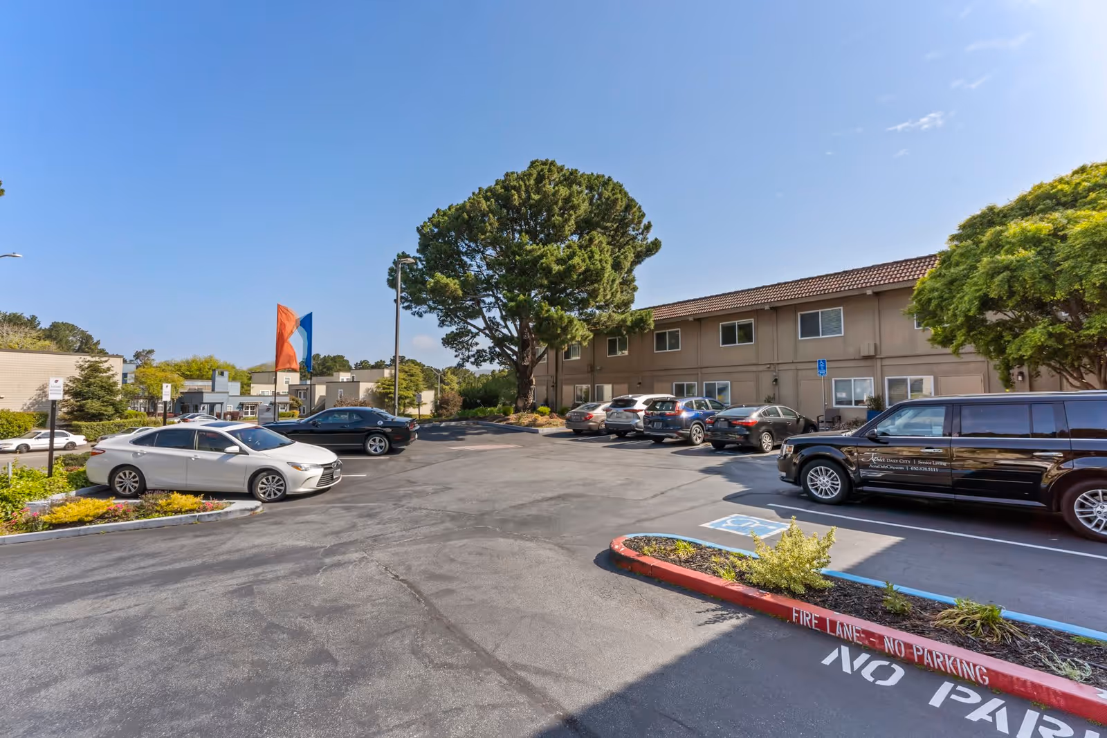 Parking lot outside a two-story senior living facility building with several parked cars, trees, and a clear blue sky.
