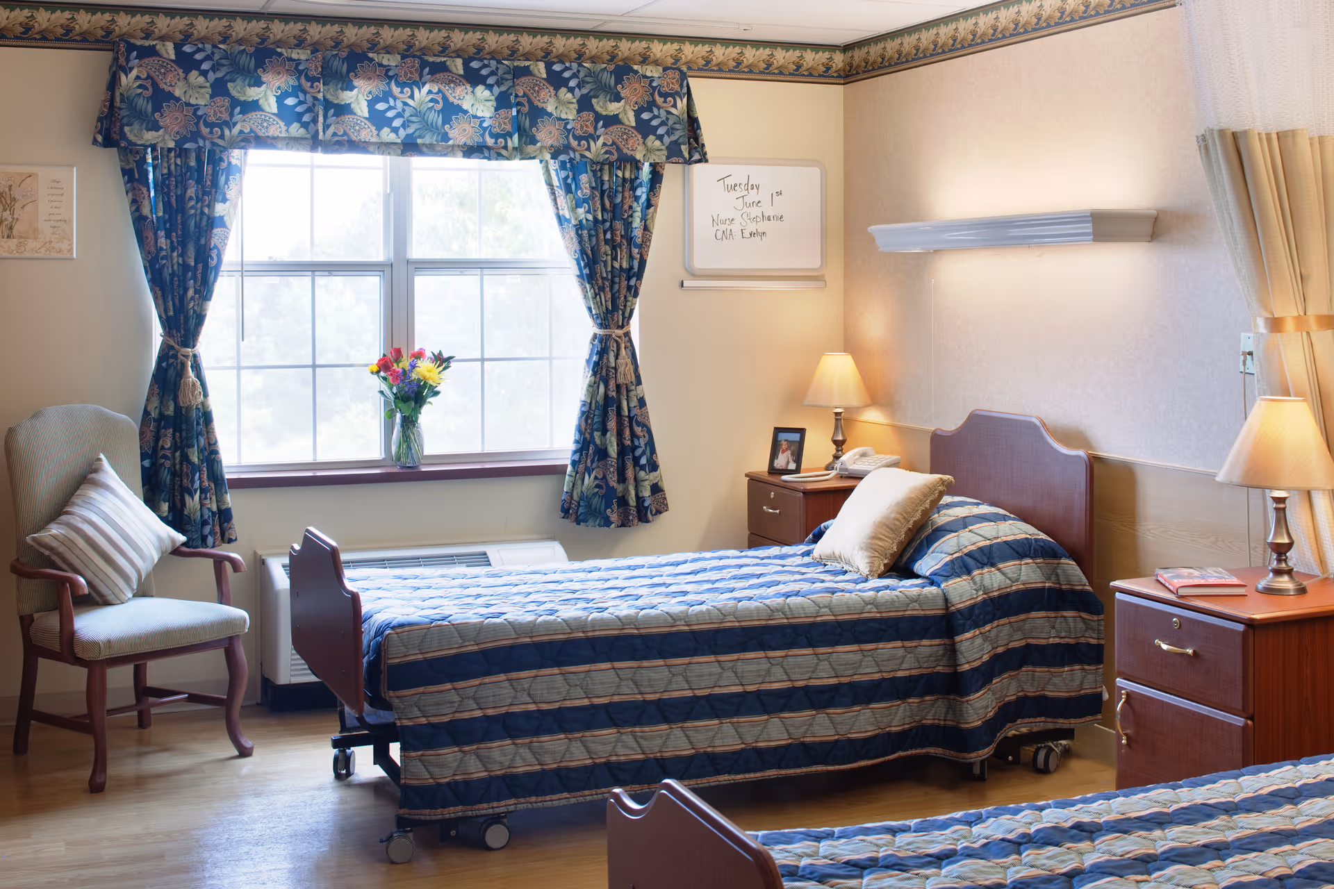 A well-lit bedroom in a senior living facility with two single beds covered in blue patterned bedspreads. There is a wooden nightstand with a lamp and a telephone between the beds. A window with floral curtains lets in natural light, and a vase with colorful flowers is placed on the windowsill. A cushioned armchair with a striped pillow is positioned near the window. A whiteboard on the wall displays a note about nurse Stephanie and CNA Evelyn.