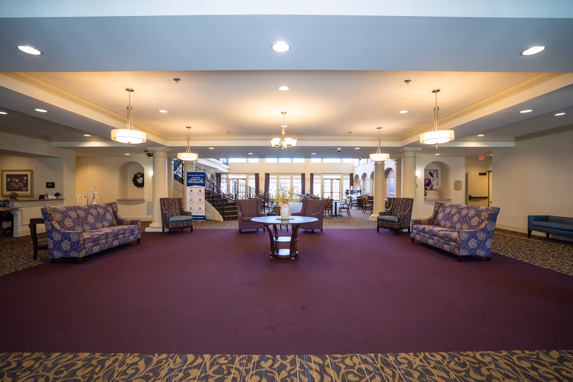 Spacious senior living lobby with purple carpet, patterned sofas, a central round table, and overhead pendant lights.