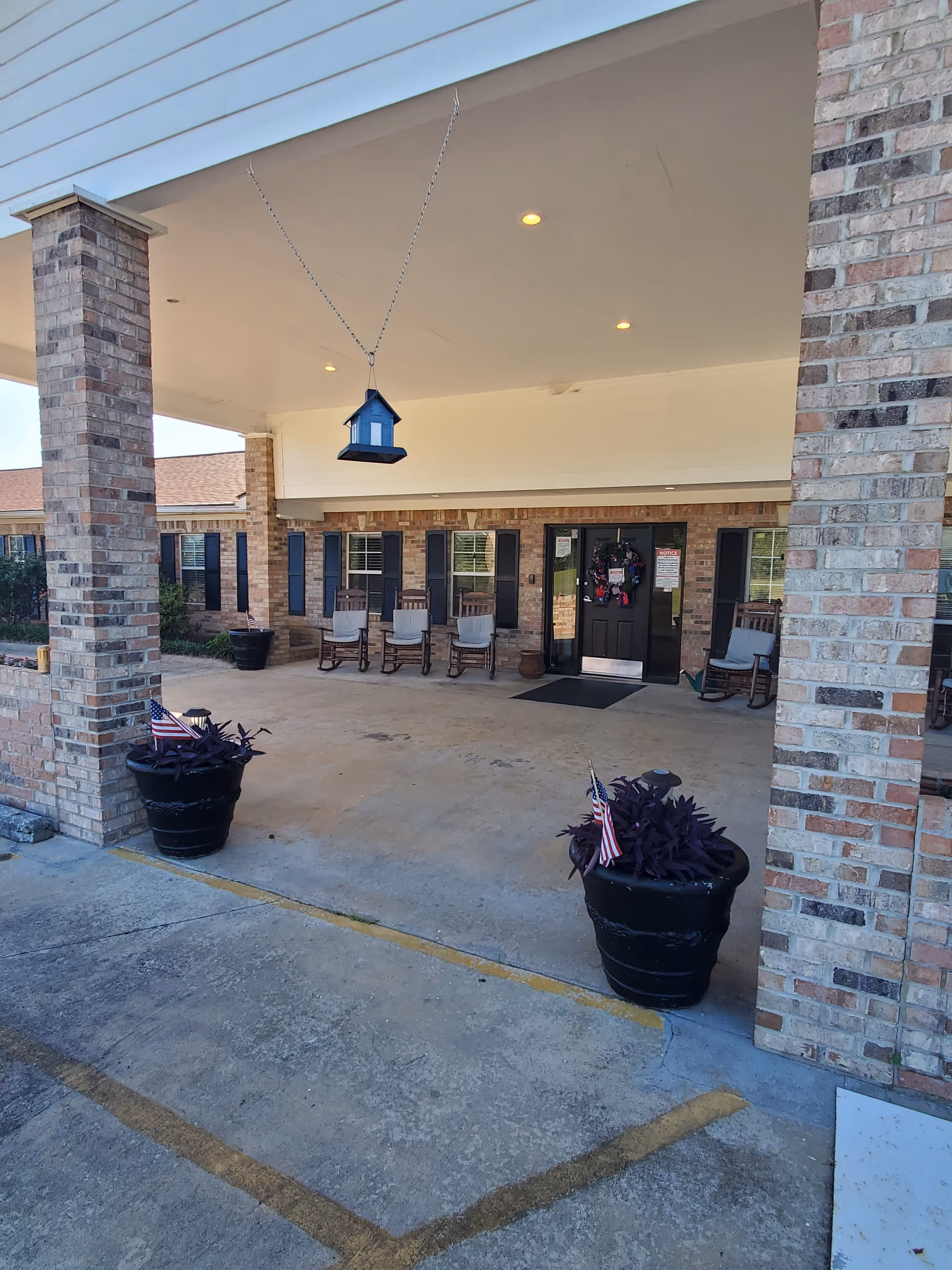 Covered entrance area of a brick building with rocking chairs lined up against the wall near the door. Two large black planters with purple plants and small American flags are placed on either side of the entrance. A small blue birdhouse hangs from the ceiling. The entrance door has a wreath and some notices posted on it.