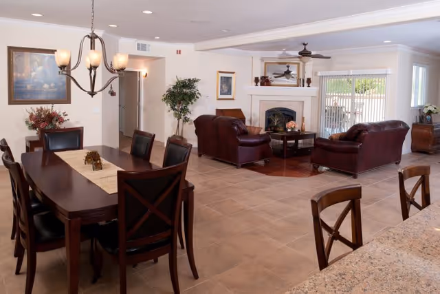 A spacious and well-lit common area in a senior living facility featuring a dark wooden dining table with six chairs on the left, a cozy living room area with two brown leather sofas and a coffee table centered around a fireplace, a ceiling fan, and large sliding glass doors leading to an outdoor space. The room has tiled floors, light-colored walls, and decorative elements including framed artwork, plants, and a chandelier above the dining table.