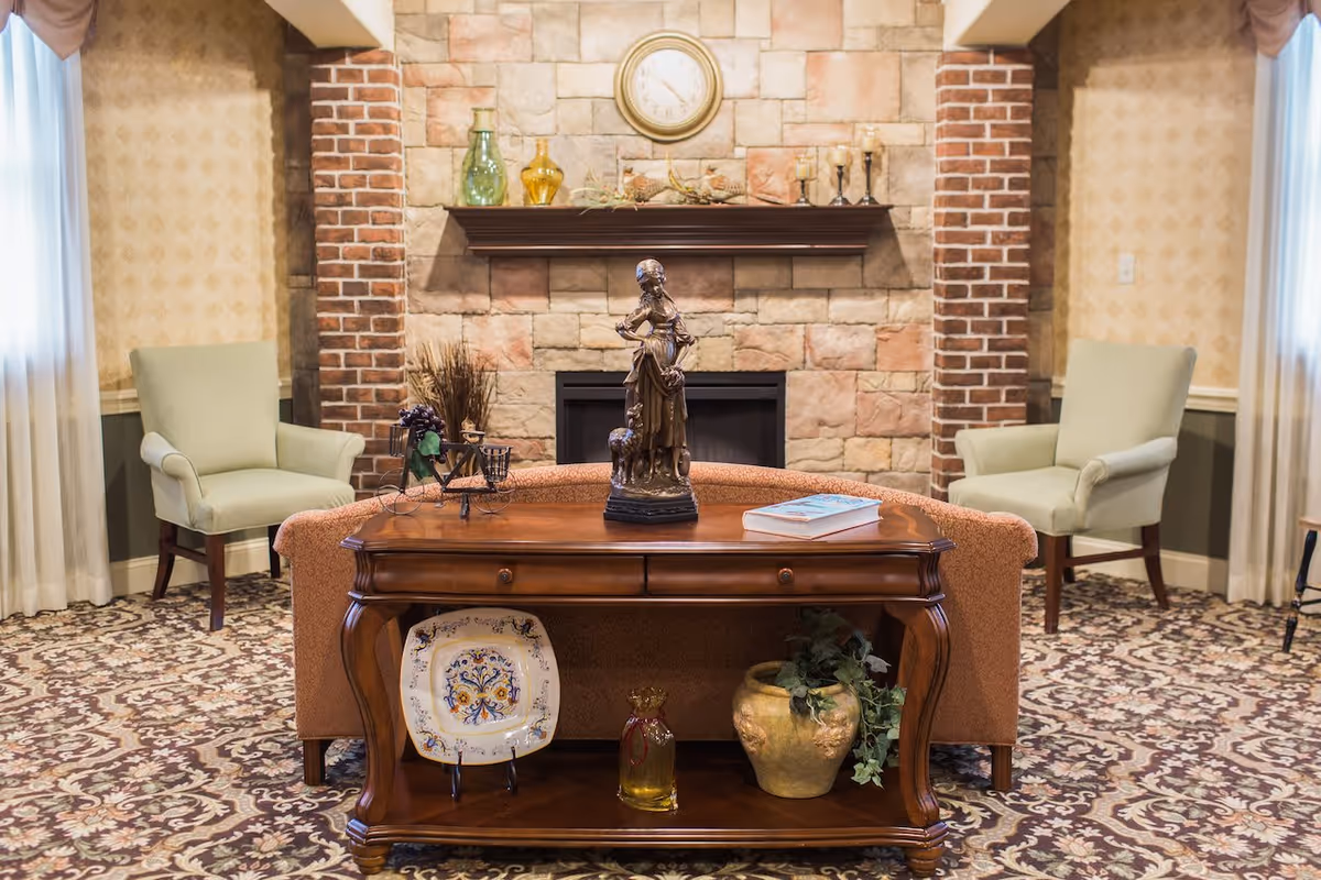 A cozy lobby area featuring a wooden table with decorative items, a statue, and a patterned rug. Two green armchairs are positioned beside a brick fireplace, creating a warm atmosphere.