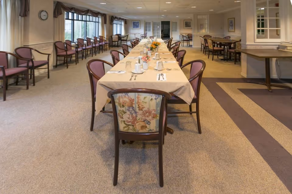 A large dining room with a long table covered with a beige tablecloth, set with cups, glasses, and utensils. The chairs around the table have floral upholstery on the backrests. Additional chairs line the walls and there are more tables and chairs in the background. The room has carpeted floors and large windows on one side.