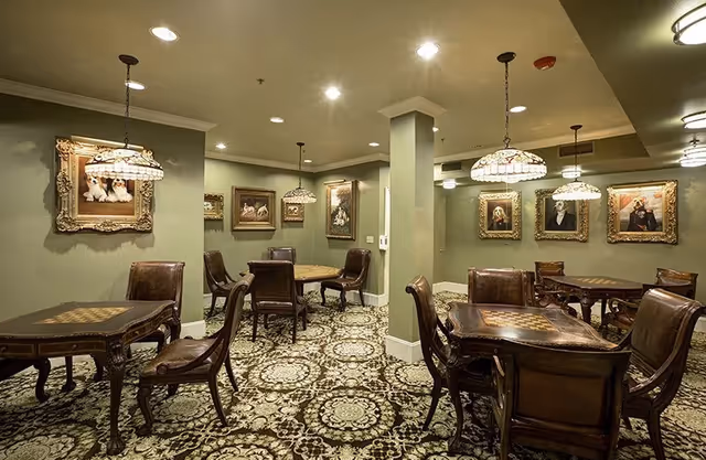 An elegant common room with wooden game tables and leather chairs beneath stained-glass pendant lamps, ornate paintings on sage-green walls, and a patterned carpet.