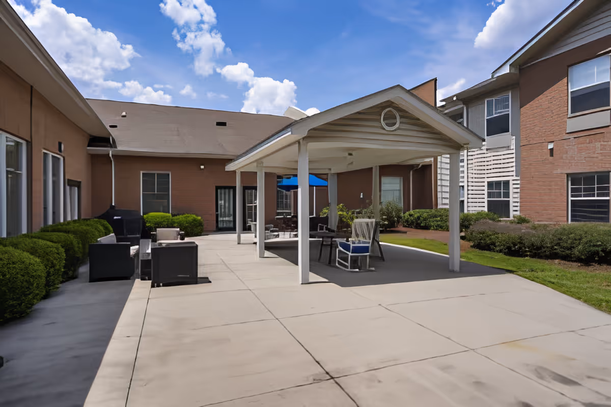 Outdoor patio area at TerraBella Windsor Lake with a covered seating area featuring chairs and a table, surrounded by bushes and adjacent to the building under a partly cloudy blue sky.