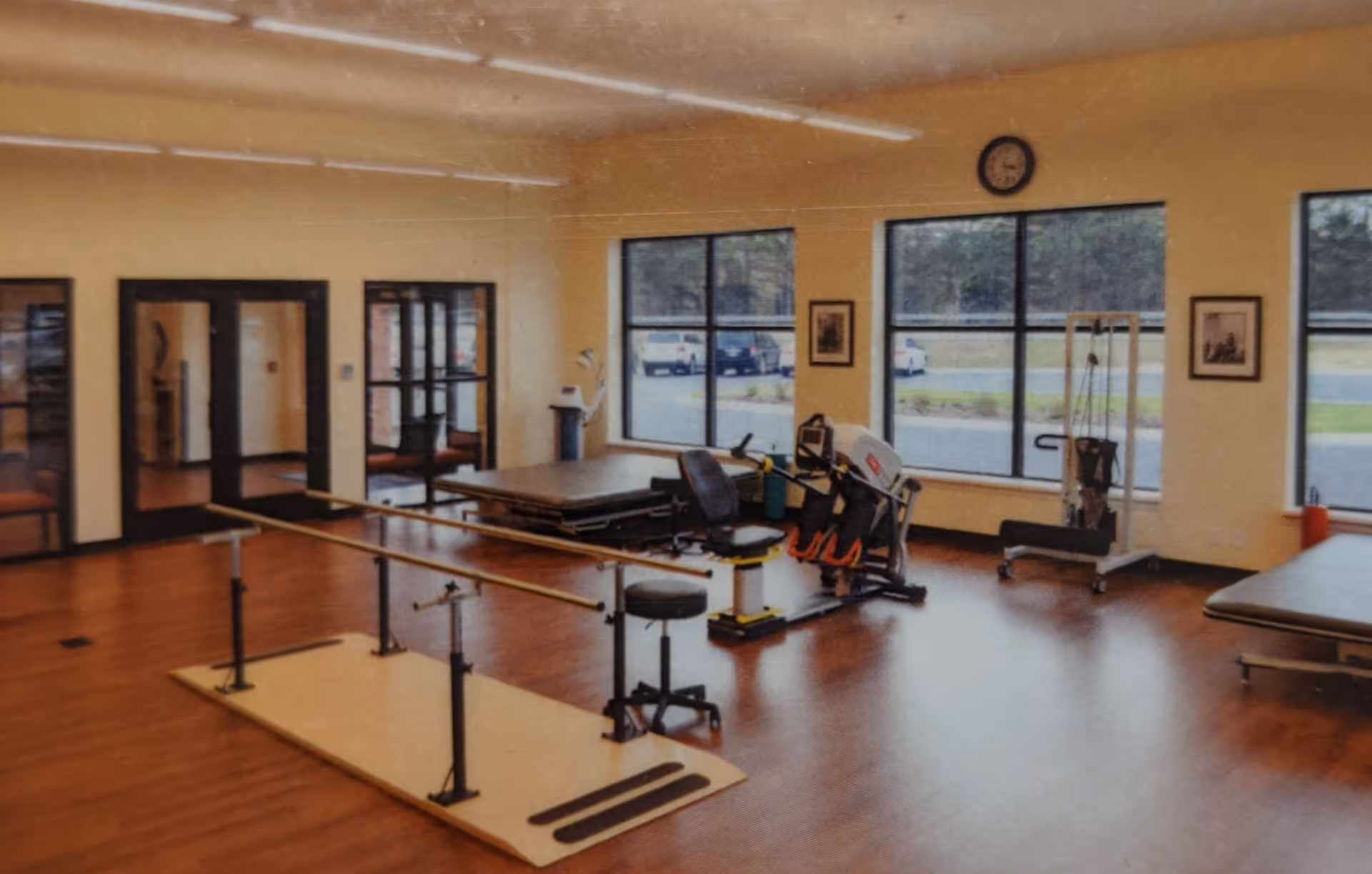 A spacious rehabilitation or physical therapy room with wooden flooring, parallel bars for walking exercises, exercise equipment, therapy tables, large windows letting in natural light, and a clock on the wall.