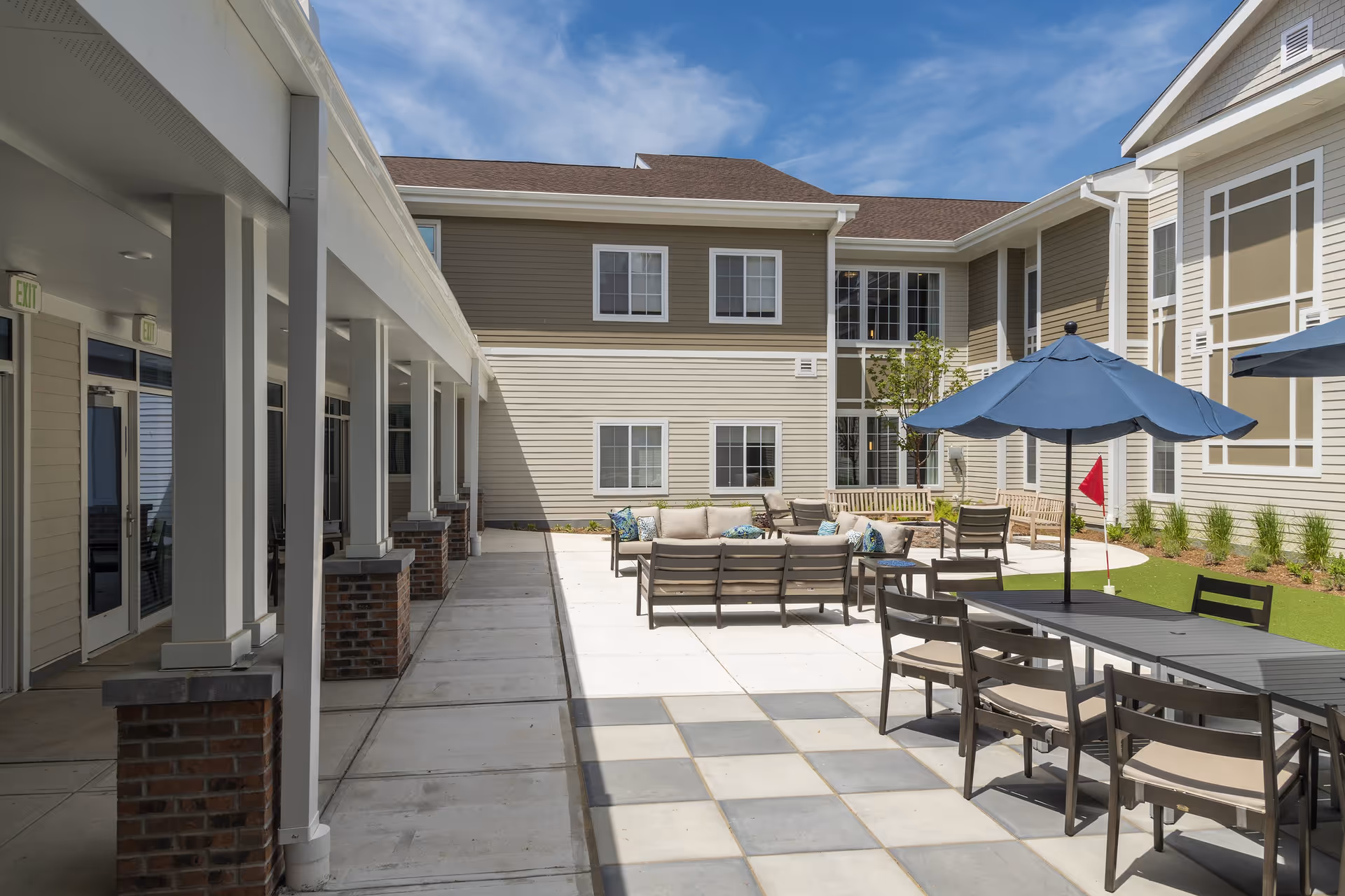 Outdoor courtyard area at Alto Evergreen Walk featuring patio seating with cushioned chairs and sofas, tables with blue umbrellas, a putting green with a red flag, and a two-story building with beige siding and multiple windows under a blue sky.