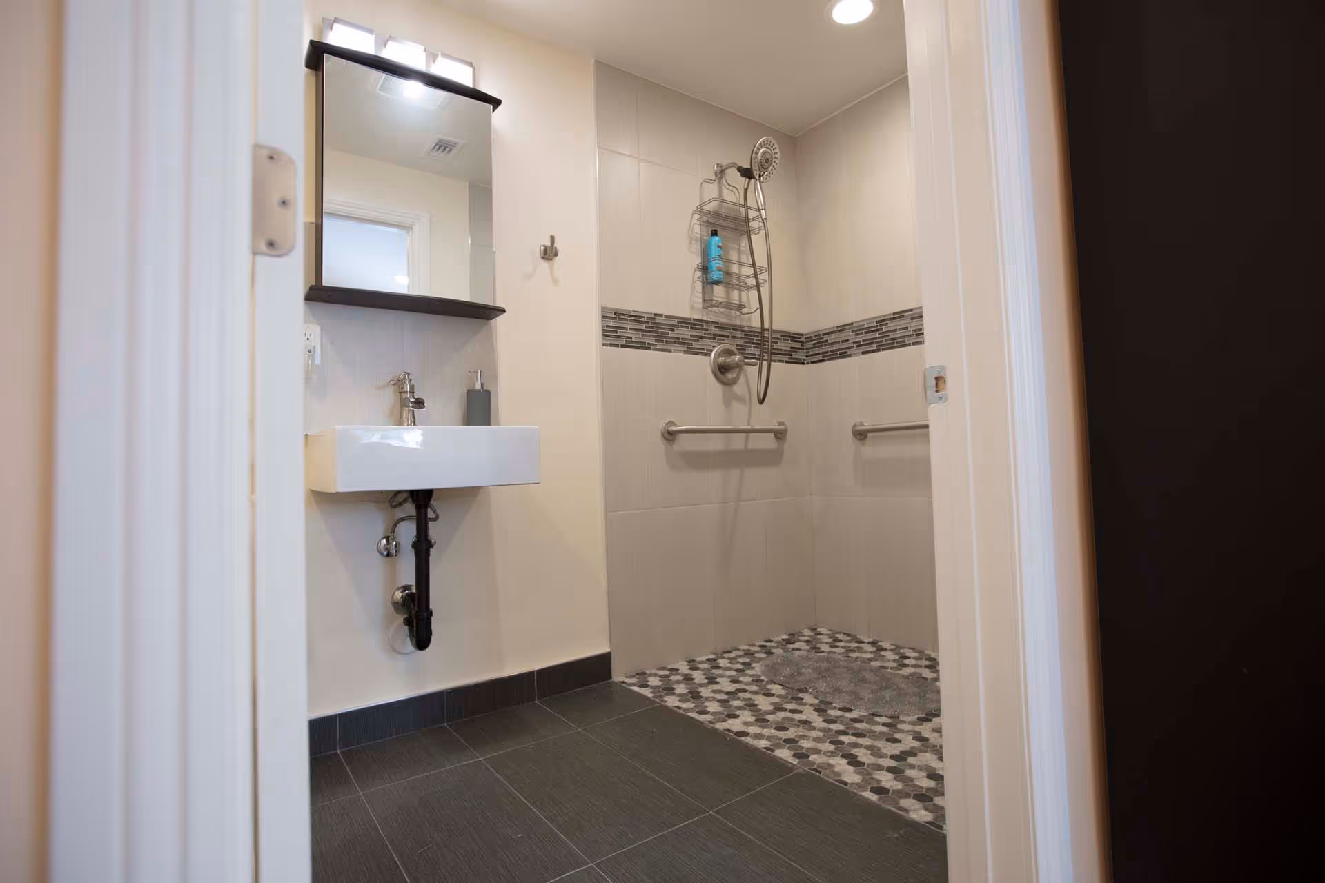 A modern bathroom with a walk-in shower featuring a handheld showerhead and a tiled floor with hexagonal patterns. The shower walls have a horizontal decorative tile strip. There are two grab bars installed on the shower walls. A small white sink with a mirror above it is mounted on the wall outside the shower area.
