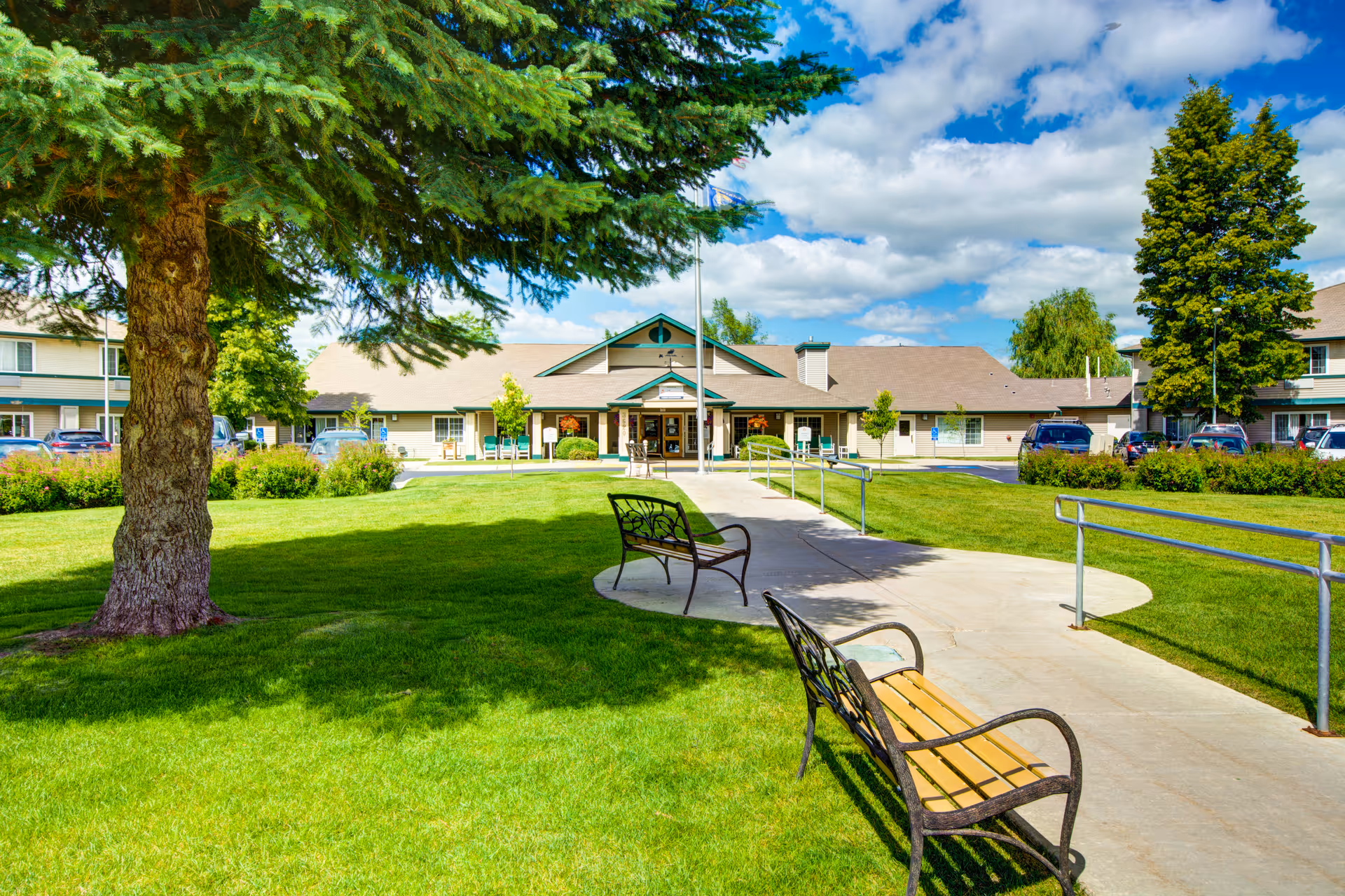 Outdoor view of Missoula Valley Senior & Assisted Living facility showing a well-maintained lawn with two benches along a curved concrete pathway, large trees providing shade, and the building entrance in the background under a partly cloudy blue sky.
