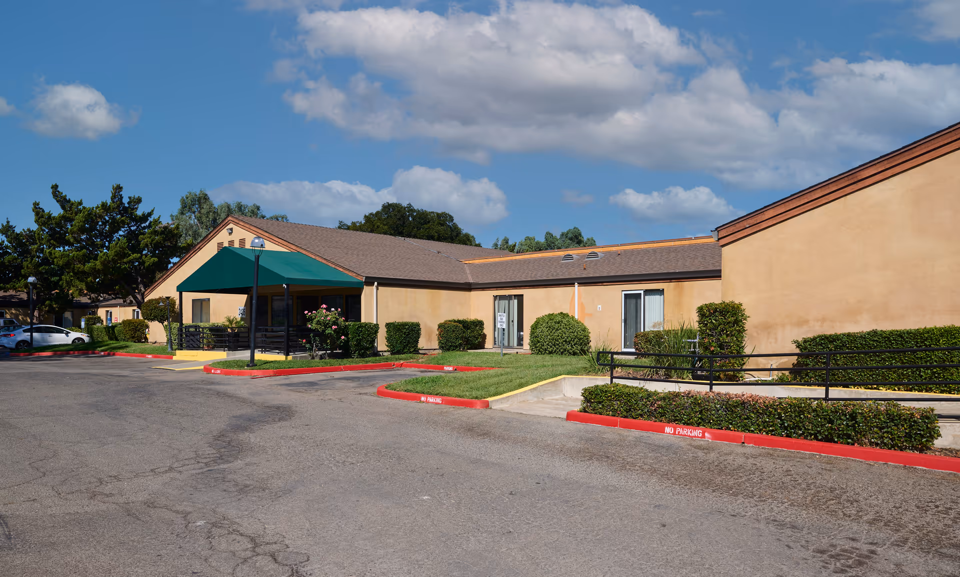 Exterior view of Wagner Heights Residential facility showing a single-story building with beige walls and a brown roof. There is a green awning over the entrance, surrounded by neatly trimmed bushes and a parking area with red curbs marked 'No Parking'. The sky is blue with scattered clouds.