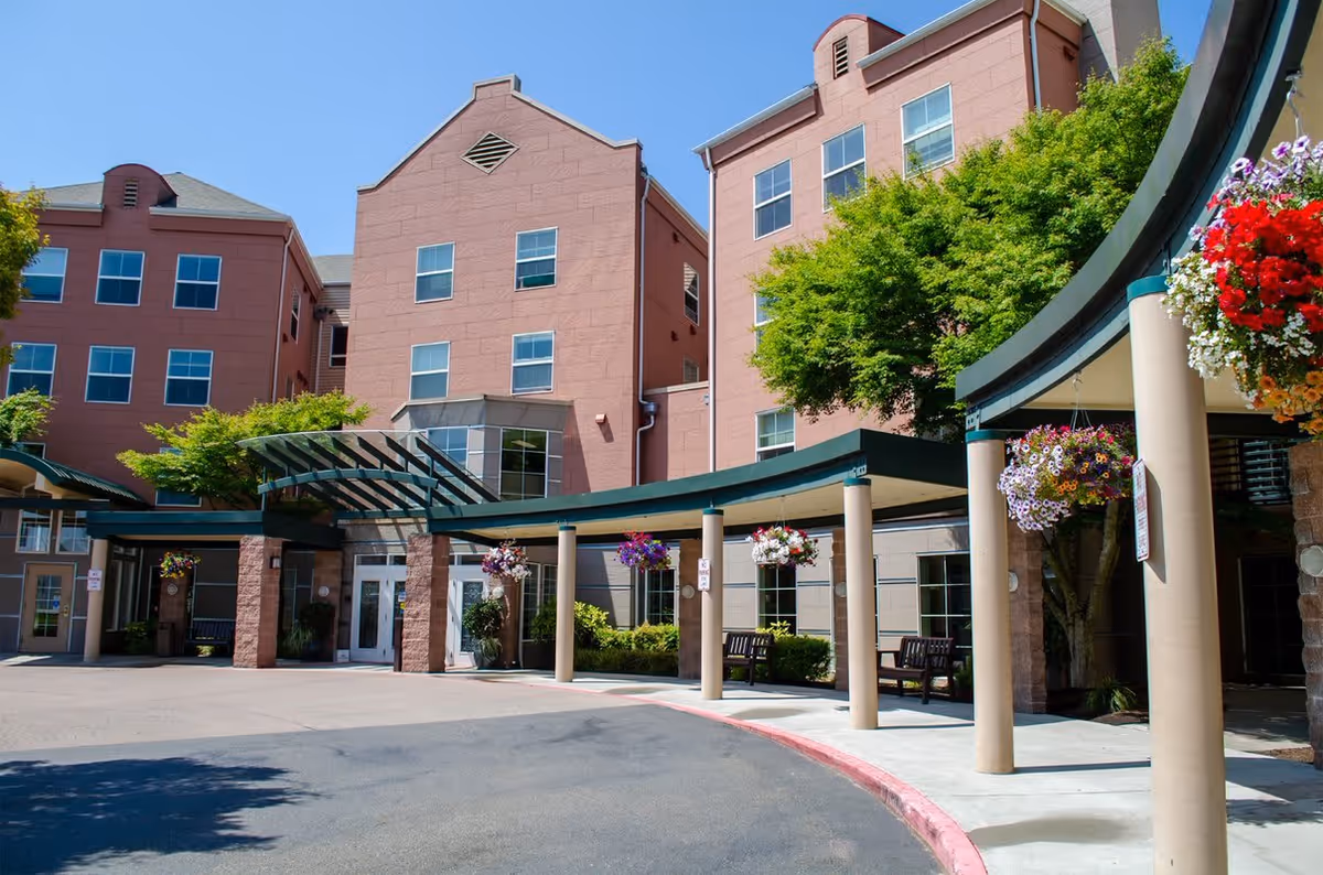 Front entrance of a multi-story pink retirement building with a covered walkway, hanging flower baskets, and benches.