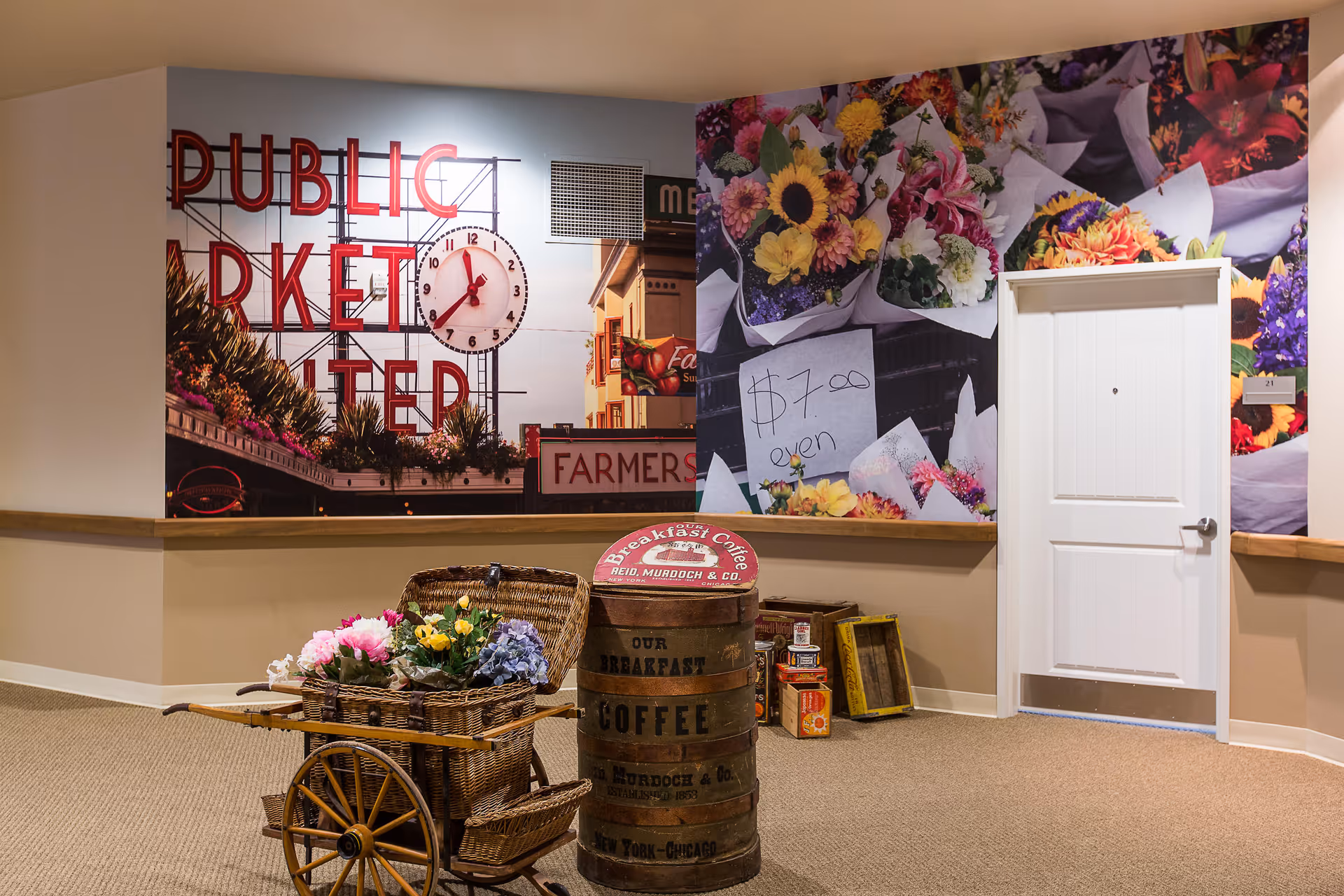 Interior hallway of a senior living facility with a large wall mural featuring a public market sign and colorful flower bouquets. In front of the mural, there is a decorative wooden cart filled with artificial flowers and a vintage barrel labeled 'Breakfast Coffee'. A white door is visible on the right side of the image.