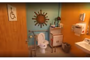 Interior view of an accessible bathroom with a toilet equipped with handrails, a sink, a soap dispenser, a small cabinet with a lamp and decorative items, a trash bin, and a wall decoration shaped like a sun. The door has a wheelchair accessibility sign.