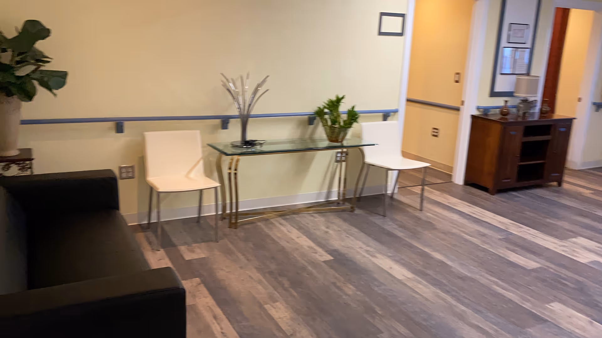 Interior view of a hallway or common area in a facility with a black couch on the left, two white chairs separated by a glass table with decorative items on top, a wooden cabinet with a lamp and framed pictures on the right, and light-colored walls with a handrail.