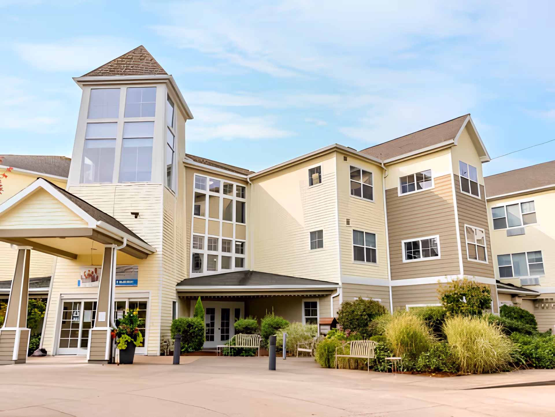 Exterior view of a multi-story senior living facility building with beige and light brown siding, large windows, a covered entrance, and landscaped greenery including bushes and benches in front.