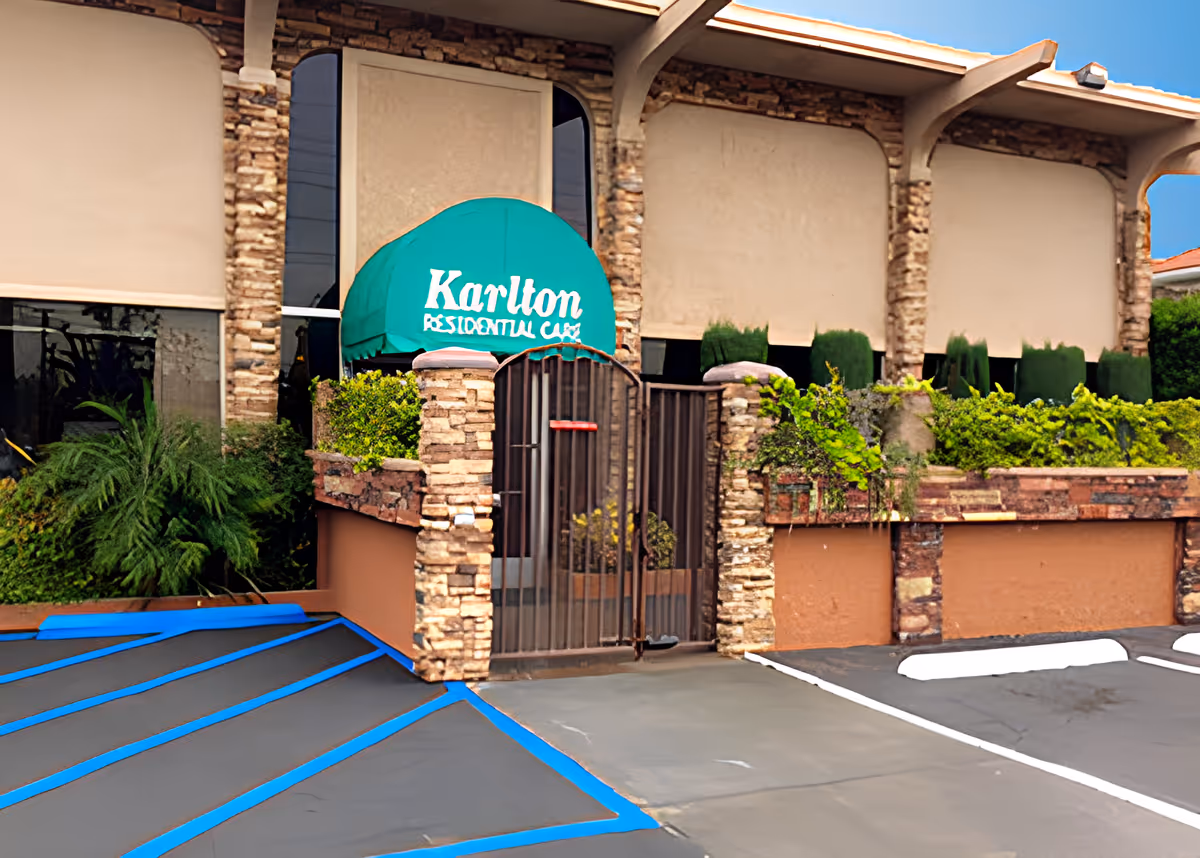Exterior view of Karlton Residential Center showing a stone and stucco building facade with a green awning displaying the facility name. There is a gated entrance with stone pillars and some greenery around the entrance. The parking area in front has marked spaces, including a handicapped parking spot.