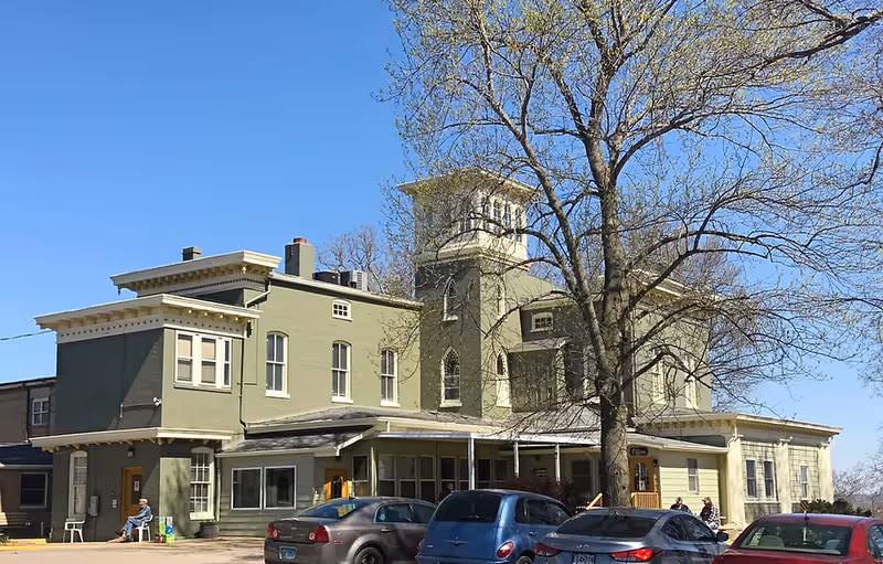 Exterior view of Marian Cliff Manor: a green, multi-story building with a small tower, cars parked in front and a large tree nearby.