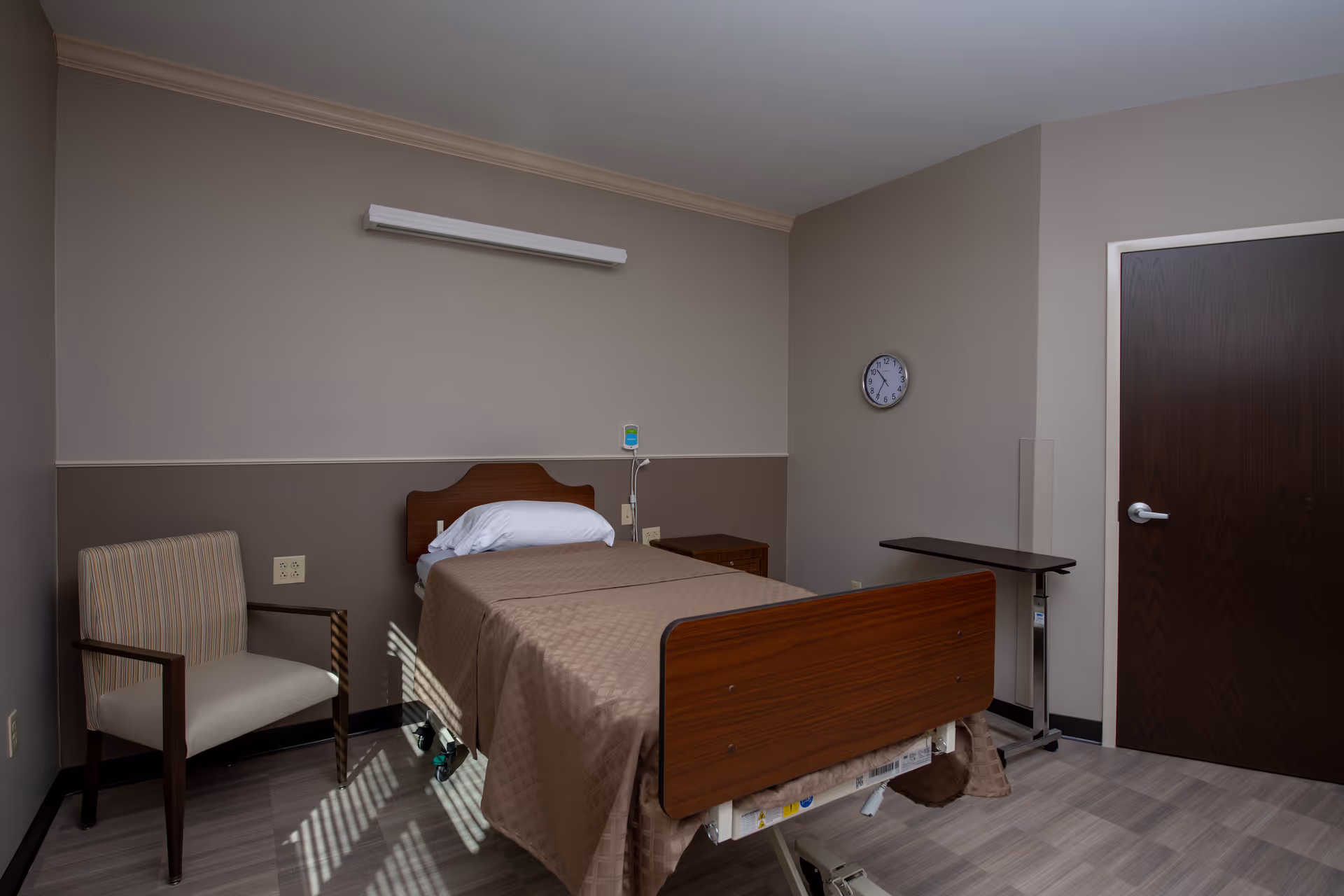 A simple healthcare facility room with a single hospital bed covered in a brown blanket, a white pillow, a wooden headboard and footboard, a striped armchair, a small wooden nightstand, a wall clock, and a dark wooden door. The walls are painted in two tones of beige and the floor has a light wood pattern.