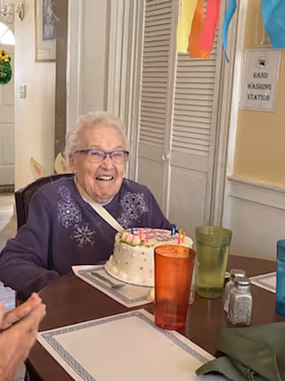 An elderly woman wearing glasses and a purple sweater sits at a dining table smiling in front of a birthday cake with lit candles. The table has placemats, colorful drinking glasses, and a salt shaker. Behind her, there is a sign on the wall that reads 'HAND WASHING STATION' and colorful decorations hanging.