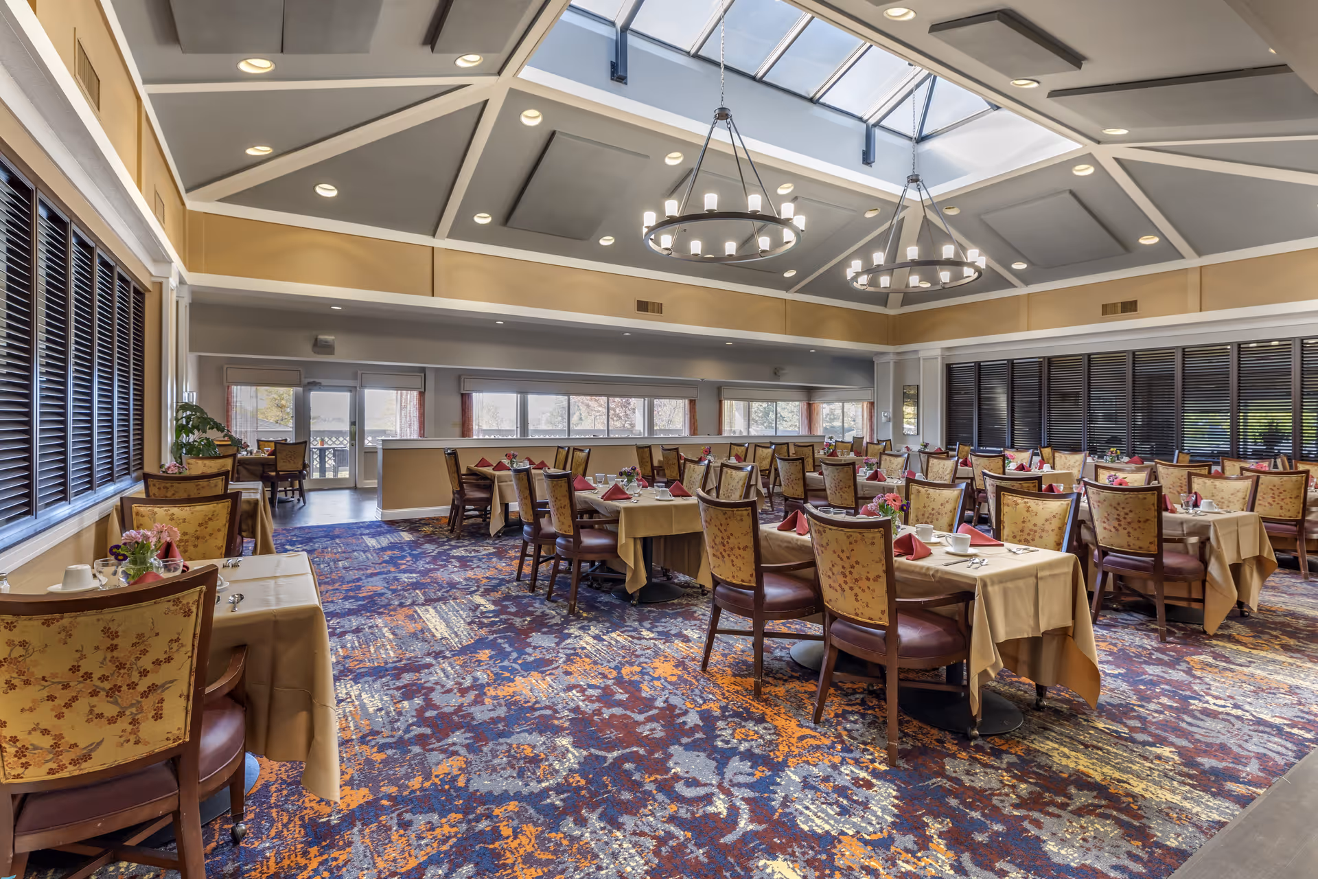 A spacious dining room with multiple tables covered in beige tablecloths, each set with cups, plates, and red folded napkins. The chairs have floral upholstery and wooden frames. The room features a colorful patterned carpet, large windows with dark blinds, and two large circular chandeliers hanging from a high ceiling with skylights.