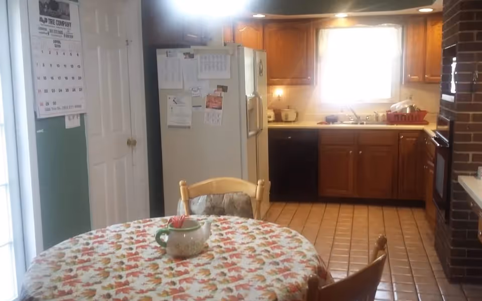 Small kitchen with wooden cabinets, a refrigerator and a round dining table with a floral tablecloth in the foreground.
