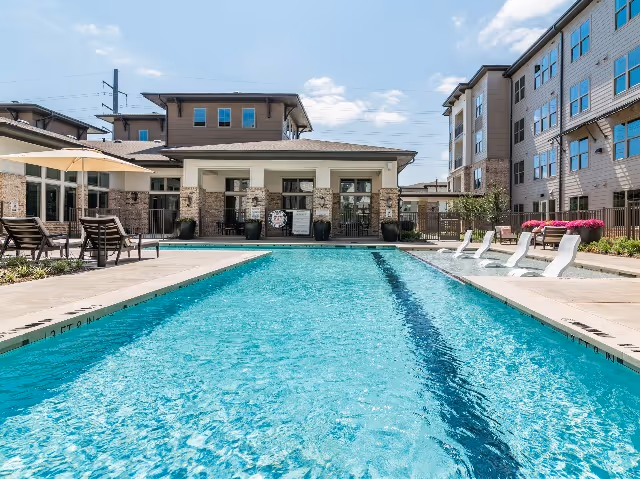 Outdoor swimming pool with clear blue water surrounded by lounge chairs and umbrellas, with a multi-story residential building and a clubhouse in the background under a partly cloudy sky.