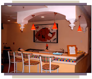 Interior view of a seating area with a counter and four bar stools with plaid cushions. The counter has a colorful tiled edge and is decorated with small model cars and a framed note. Above the counter, there are four red pendant lights hanging from a white ceiling structure. A framed artwork featuring a red jacket is mounted on the beige wall behind the counter.