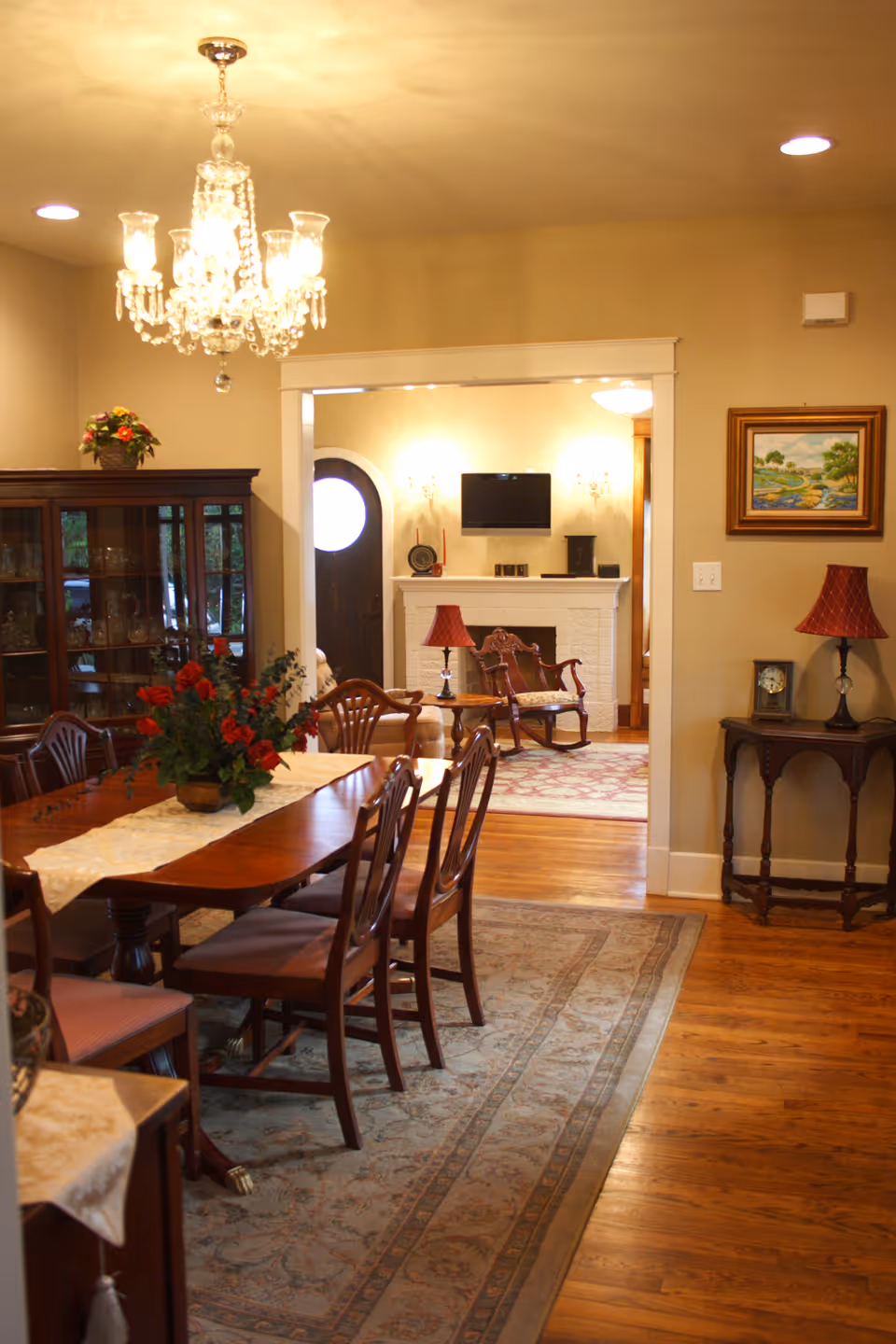 Dining room with a long wooden table and chairs under a crystal chandelier, leading into a living room with a fireplace and TV.