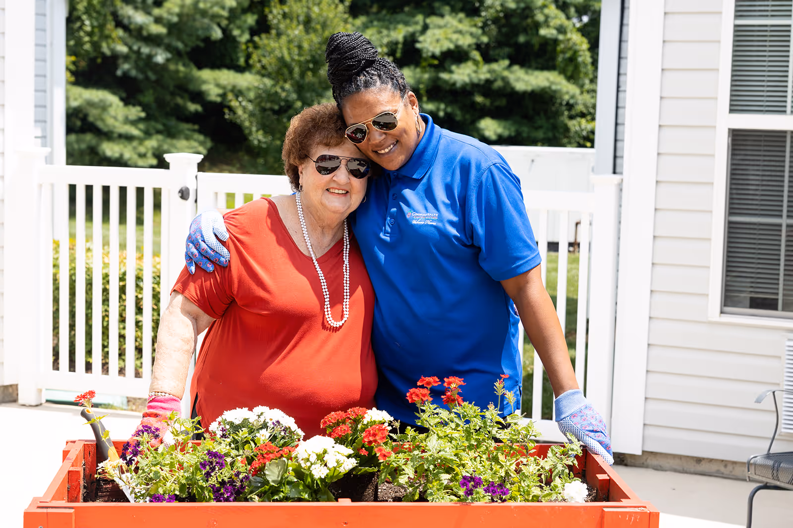 Two women smiling and posing together outdoors by a raised garden bed filled with colorful flowers. One woman is elderly wearing a red shirt and sunglasses, and the other is younger wearing a blue polo shirt and gardening gloves. They are standing in front of a white fence and a building with siding and windows.