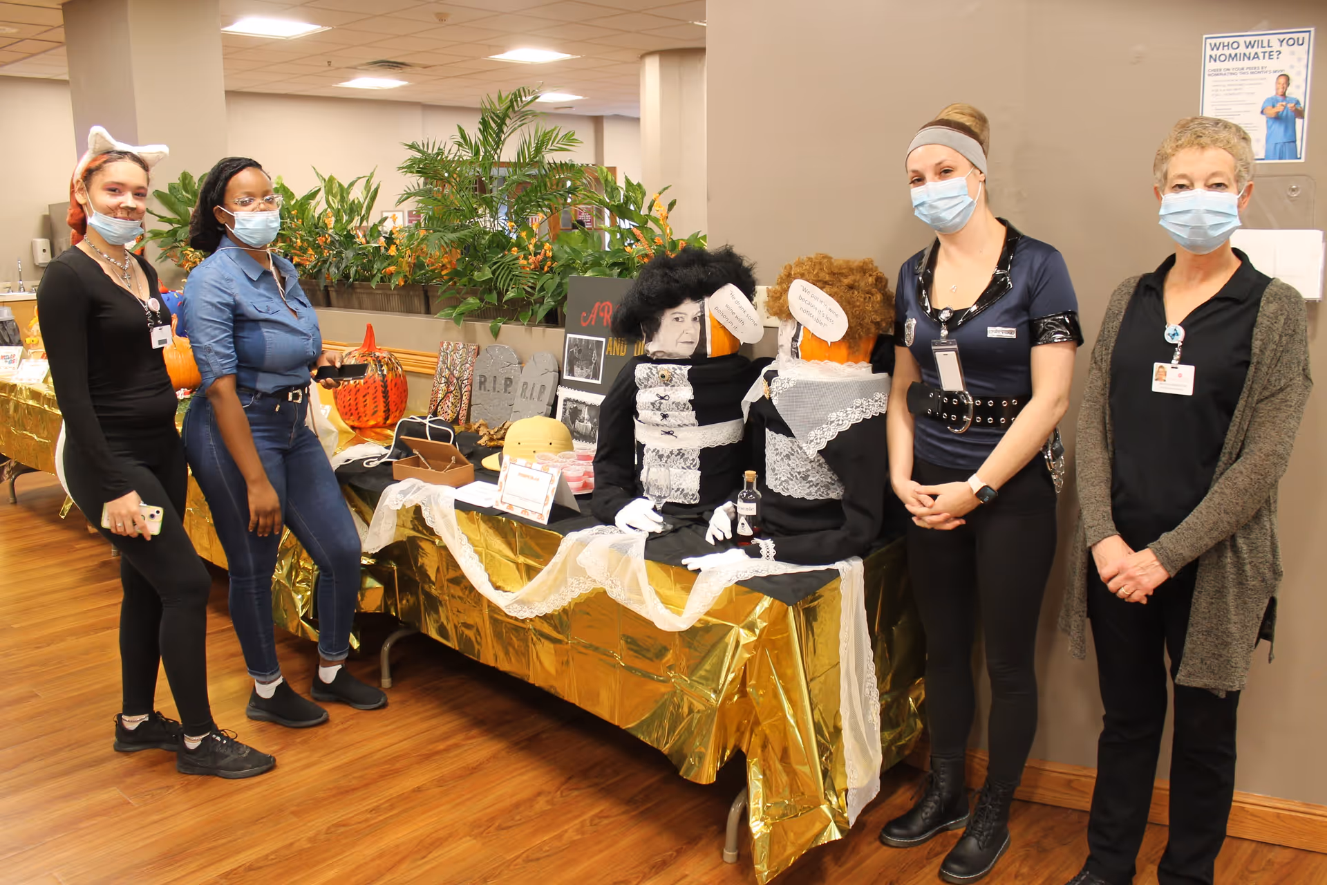 Four women wearing face masks stand next to a decorated table with Halloween-themed items including two dressed pumpkins with wigs and speech bubbles, and other festive decorations in an indoor setting with plants in the background.