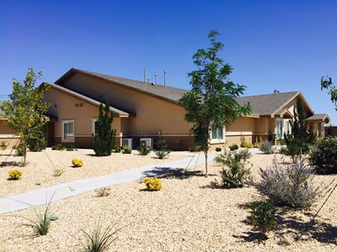 Single-story beige building with a sloped roof surrounded by a desert-style landscape featuring small trees, shrubs, and gravel ground cover under a clear blue sky.