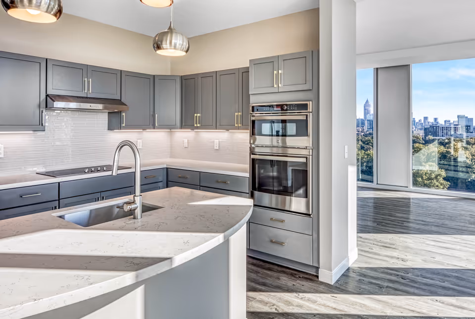 Modern kitchen with gray cabinets, stainless steel appliances including a double oven and stovetop, a curved white marble countertop with an integrated sink and faucet, and large windows showing a cityscape view in the background.
