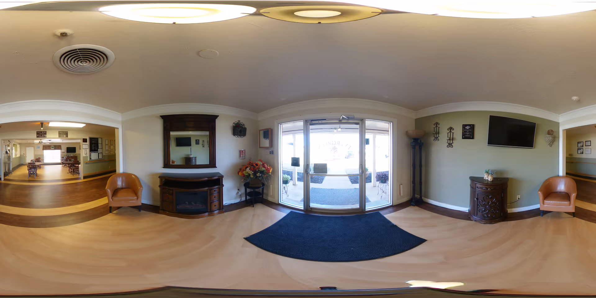 Bright reception lobby with glass entry doors, seating chairs, decorative cabinets, and a wall-mounted TV.