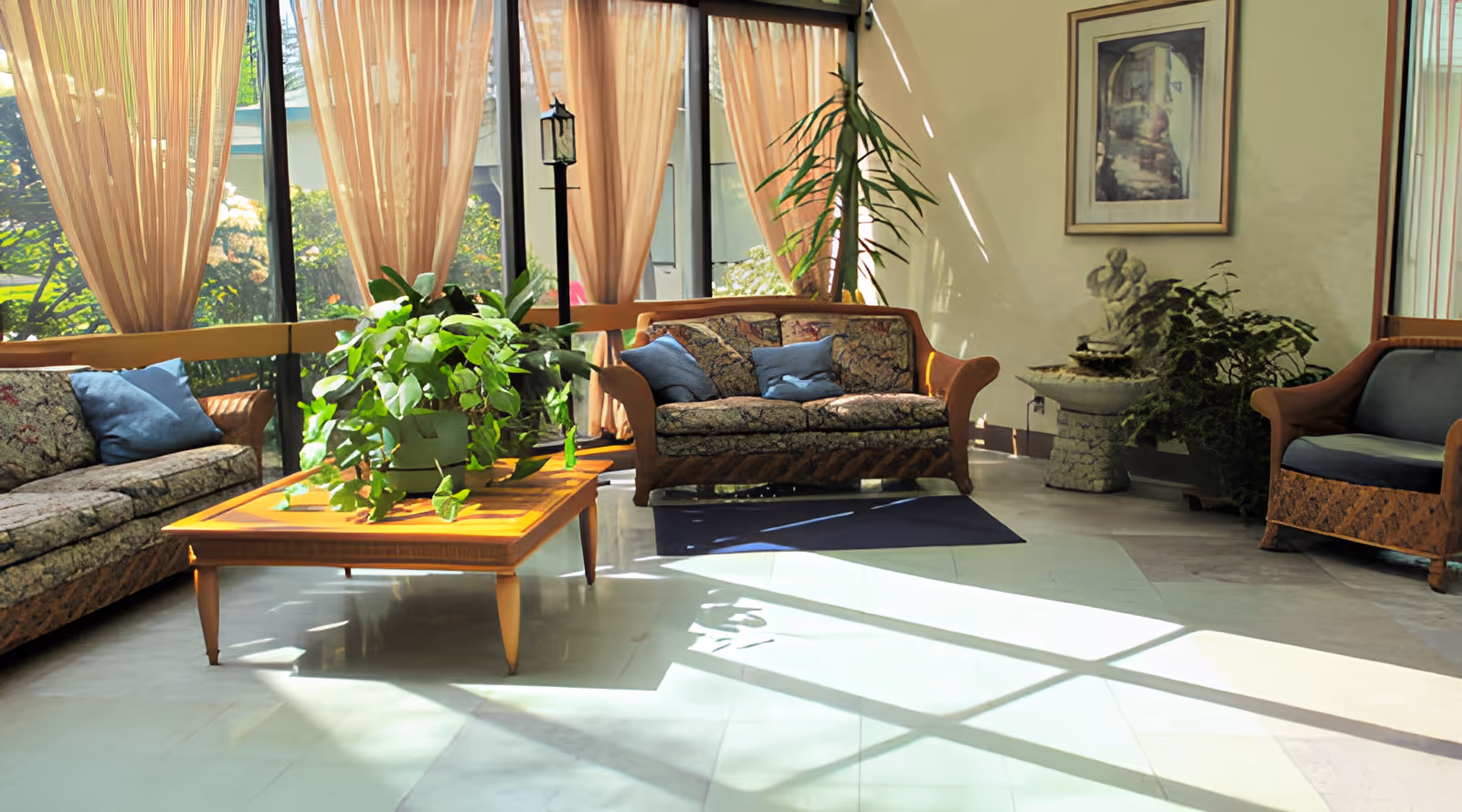 A bright and airy living room area with large windows covered by sheer curtains allowing sunlight to stream in. The room features a wooden coffee table with a green potted plant, two floral patterned sofas with blue cushions, a single armchair, a tall indoor plant, a small water fountain, and a framed picture on the wall.
