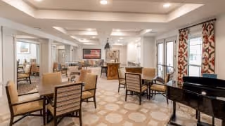 A spacious and well-lit common area in a senior living facility featuring multiple tables and chairs arranged for socializing or dining. The room has large windows with patterned curtains allowing natural light to enter. There is a piano on the right side and an American flag displayed on the far wall. The decor is warm and inviting with neutral tones and a patterned carpet.