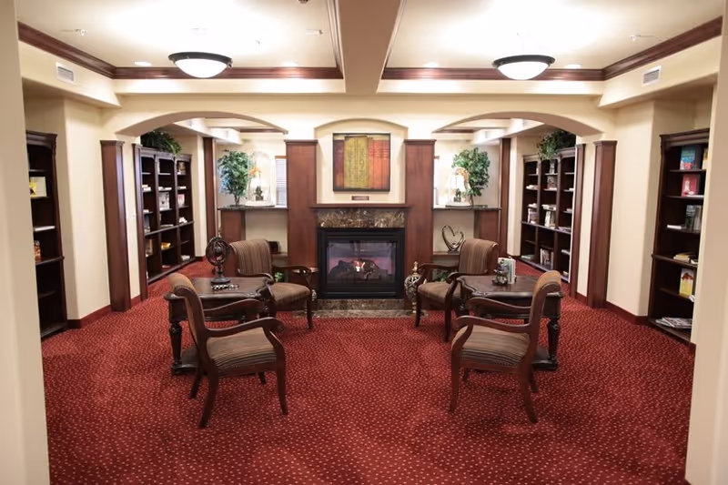 A cozy interior room with a red carpet featuring small white dots, four wooden chairs with striped upholstery arranged around two small wooden tables, a fireplace with a decorative mantle and a framed artwork above it, and bookshelves lining the walls on both sides filled with books and decorative items. There are also two potted plants and soft lighting from ceiling fixtures.