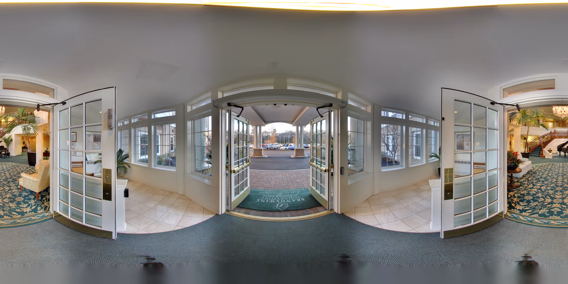 Entrance area of Brandywine Fenwick Island by Monarch showing double glass doors opening to a covered outdoor drop-off area with a green welcome mat. Inside, there is a carpeted lobby with patterned rugs, comfortable seating, plants, and a grand piano near a staircase with a chandelier above.