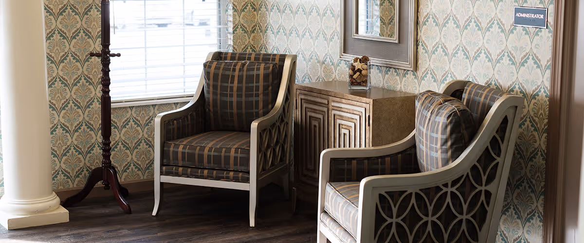 A cozy interior corner with two patterned armchairs facing each other, a small wooden cabinet between them, a tall wooden coat rack, and a window with blinds. The walls have a decorative wallpaper, and there is a sign labeled 'ADMINISTRATOR' on the wall.