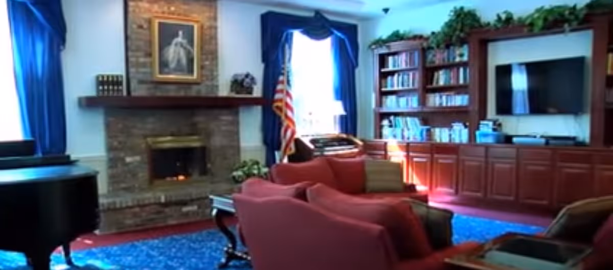 A cozy living room with red sofas, a brick fireplace with a framed picture above it, an American flag near a window with blue curtains, a grand piano, and a large wooden bookshelf with a TV mounted on the wall.