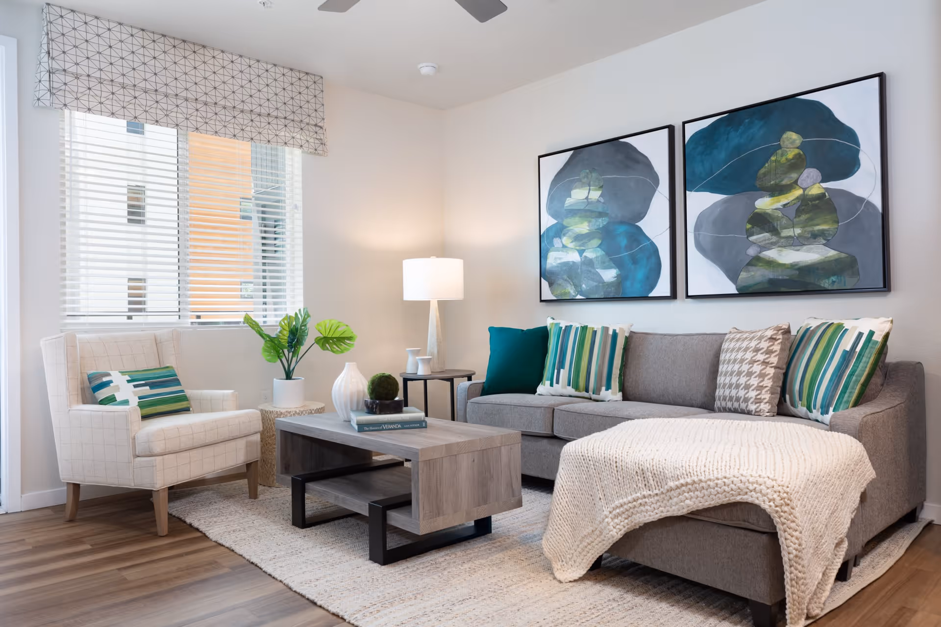 A modern living room with a gray sofa adorned with green, white, and patterned pillows, a cream knitted throw blanket draped over the sofa, a light beige armchair with a green and white striped pillow, a wooden coffee table with decorative items and books, a side table with a lamp, two abstract paintings with blue and green tones on the wall, and a window with white blinds and a patterned valance.
