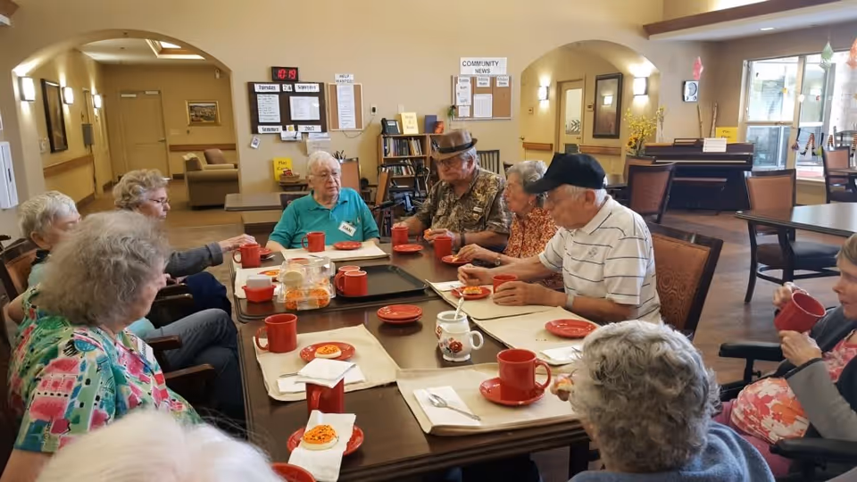 A group of elderly people sitting around a large rectangular table in a communal dining area, enjoying snacks and drinks served on red plates and mugs. The room has warm lighting, arched doorways, and a bulletin board with community news in the background.
