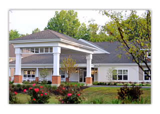 Exterior view of a single-story senior living facility building with a covered entrance supported by white columns. The building is surrounded by greenery, including bushes with red flowers and trees in the background.