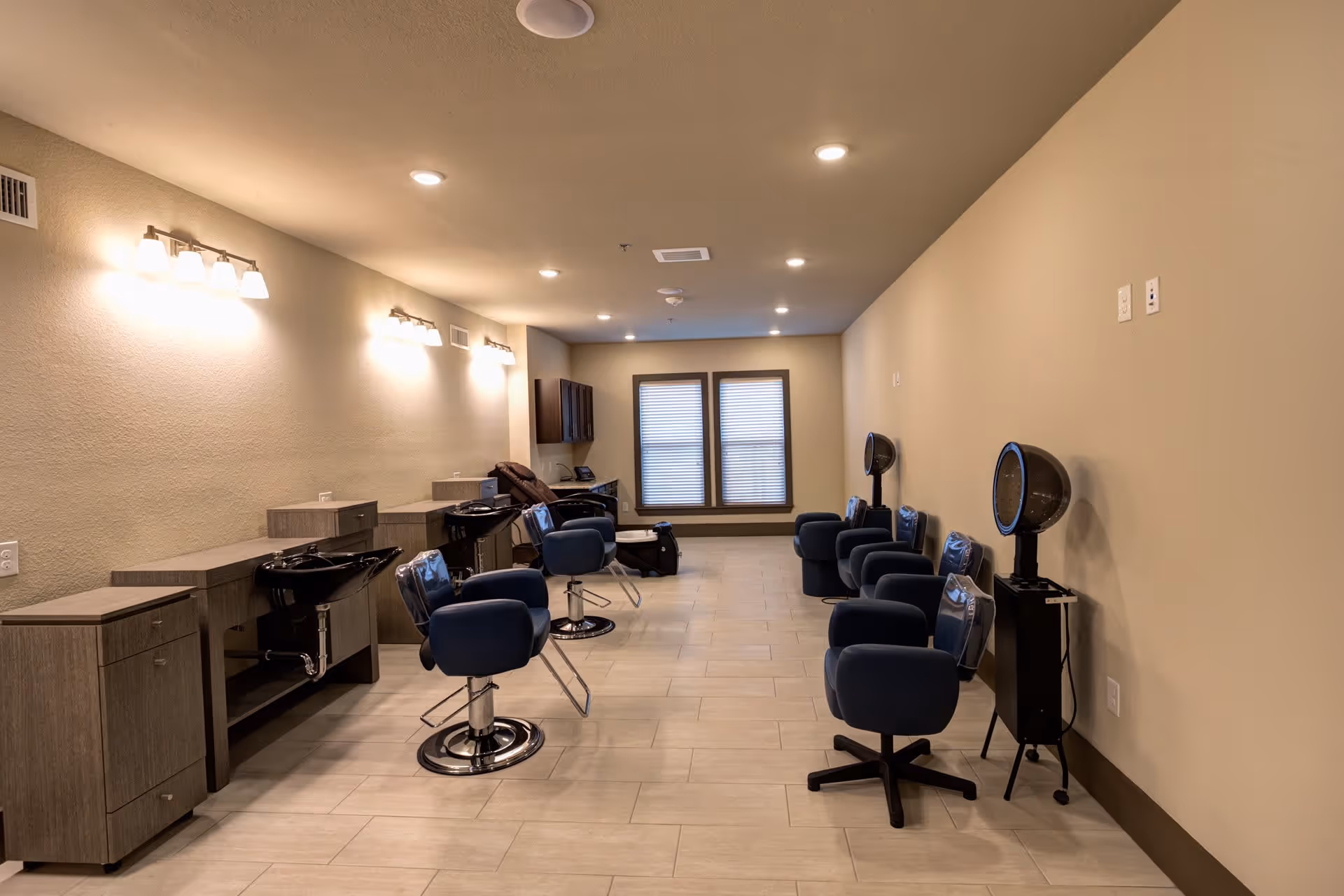Interior salon room with multiple styling chairs, hair-washing sinks, and hooded dryers arranged along a long, well-lit space.