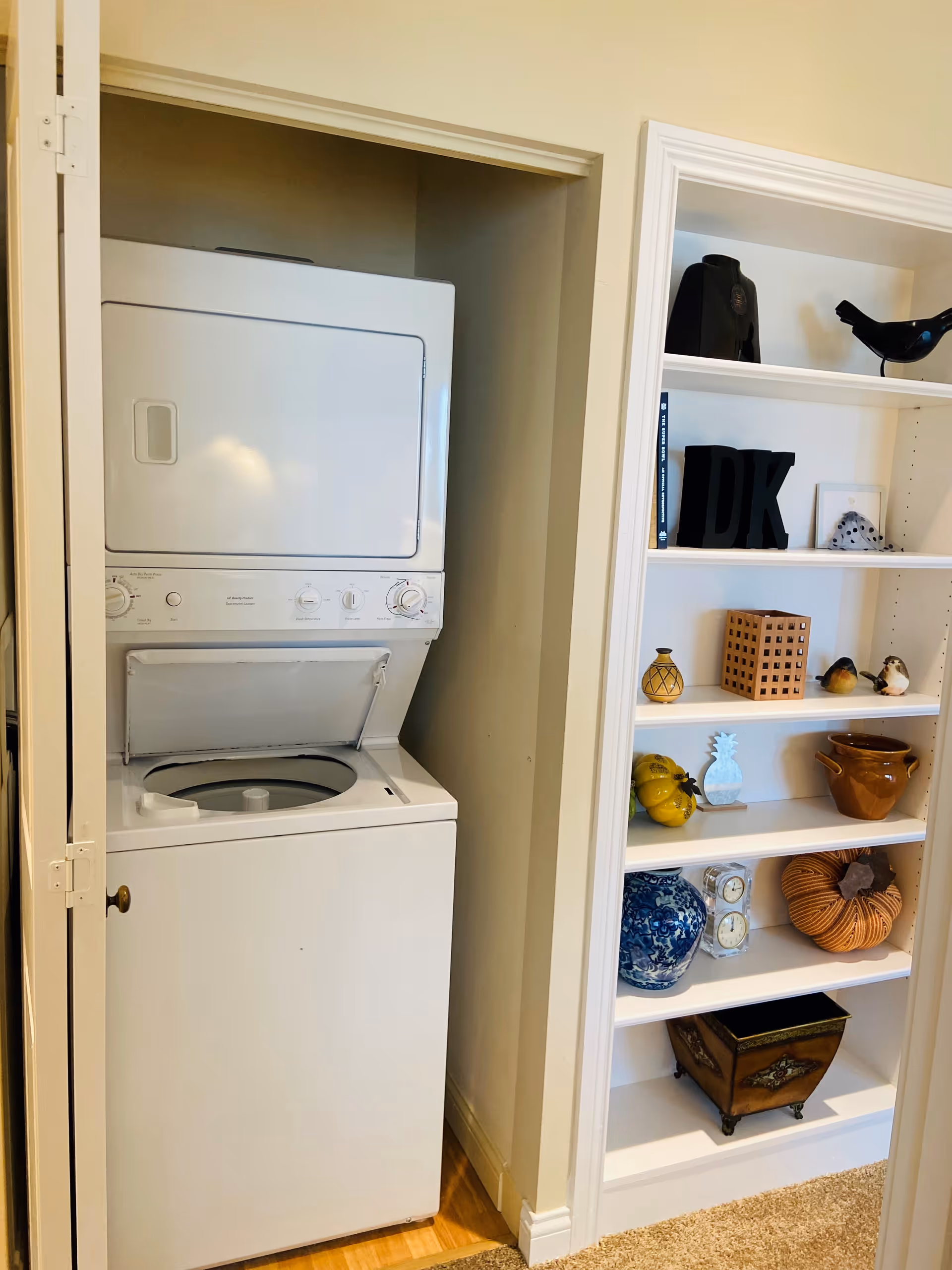 Stacked washer and dryer in a small closet next to built-in shelves filled with decorative items.