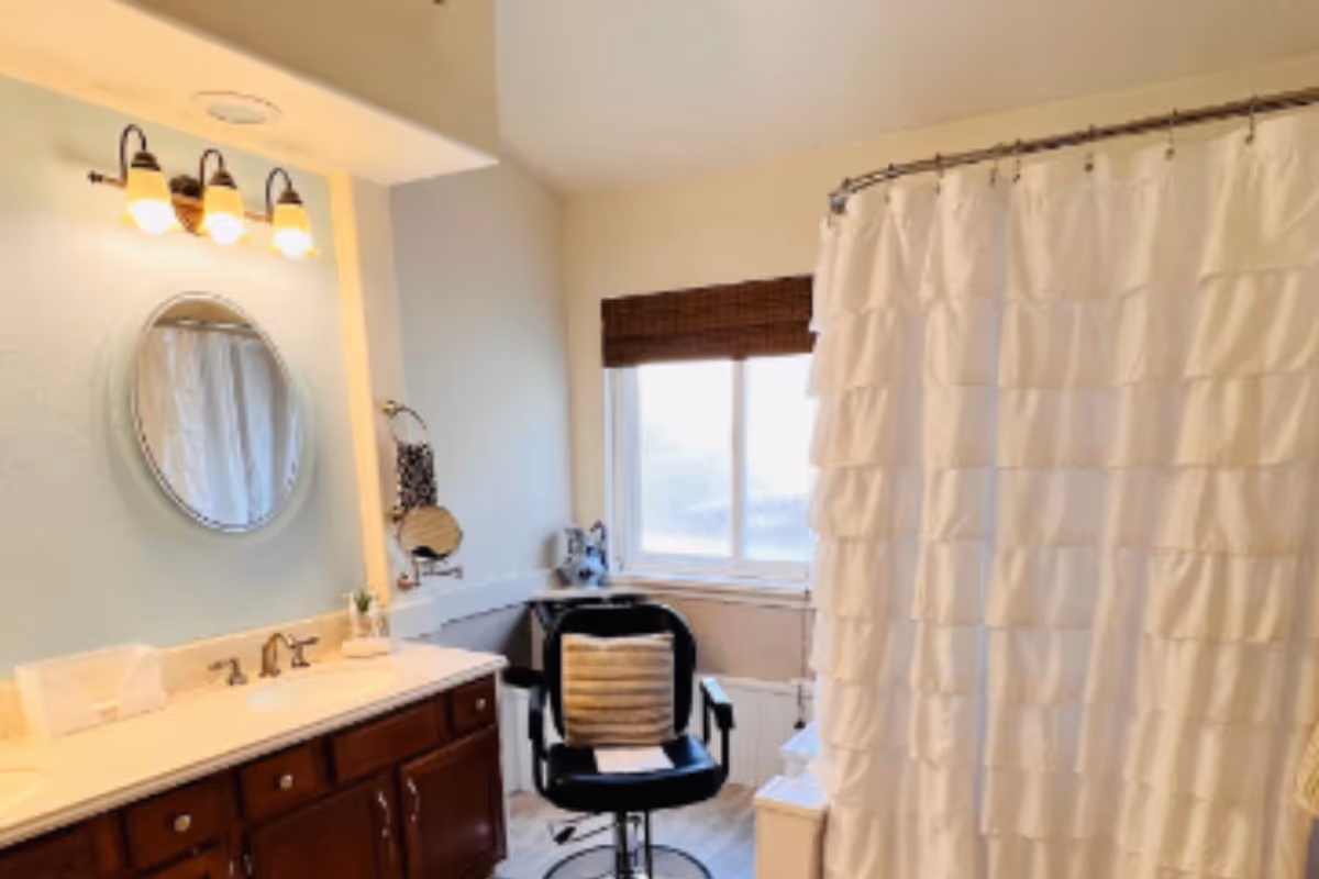 Bright bathroom with a vanity and round mirror on the left, a swivel chair by a window, and a ruffled white shower curtain on the right.