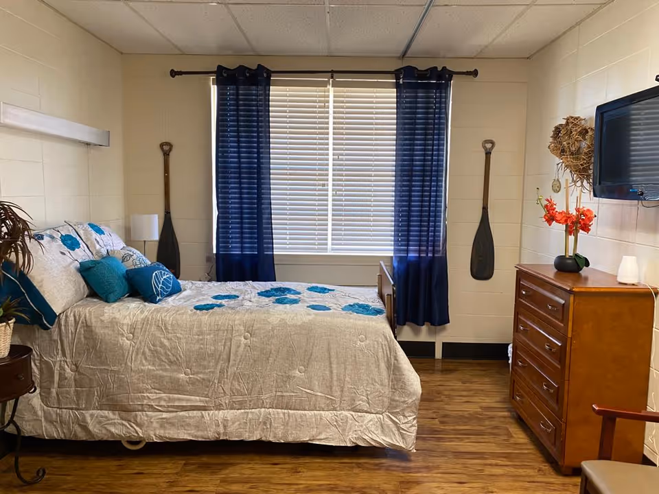 A bedroom with a bed covered in a beige bedspread with blue floral patterns and several blue pillows. The room has wooden flooring, a window with white blinds and dark blue curtains, two decorative paddles mounted on the wall on either side of the window, a wooden dresser with a vase of red flowers and a mounted TV above it, and a small side table with a plant.