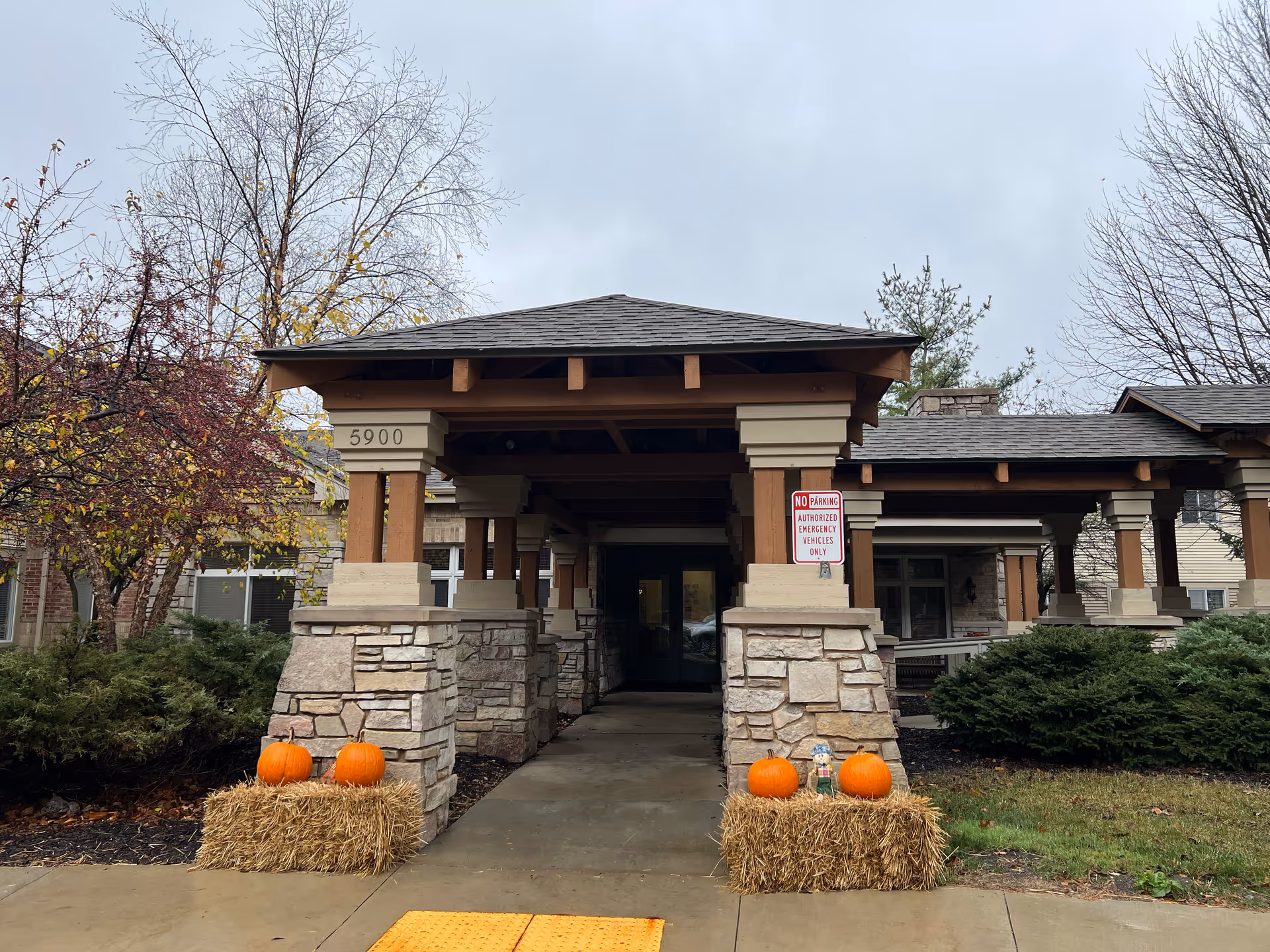 Entrance to a building with a covered walkway supported by stone and wooden pillars. There are two hay bales decorated with pumpkins and a small scarecrow figure on either side of the walkway. Trees with sparse leaves and bushes surround the entrance. The building number 5900 is visible on one pillar, and a 'No Parking Authorized Emergency Vehicles Only' sign is posted on another pillar.