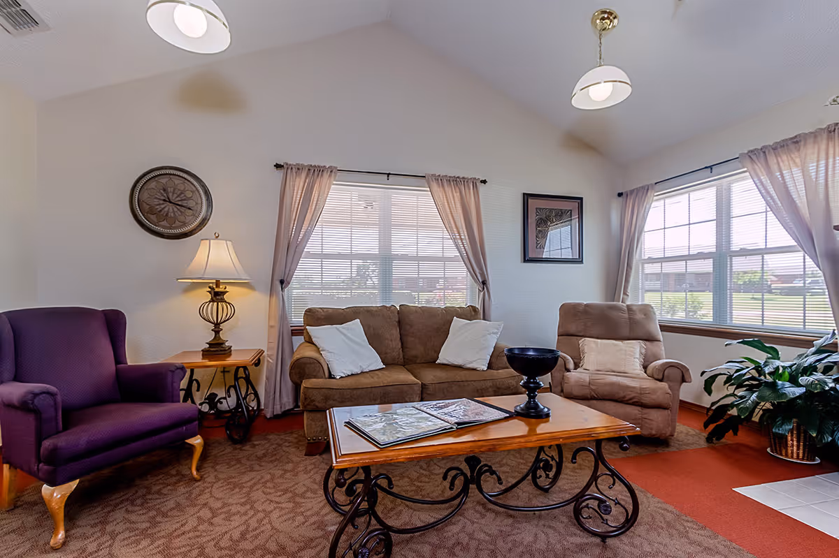 A cozy living room with a brown sofa with two white pillows, a beige recliner chair, and a purple armchair. There is a wooden coffee table with decorative wrought iron legs, holding an open book and a black decorative bowl. The room has two large windows with sheer curtains, a side table with a lamp, a wall clock, a framed picture, and a potted plant near the window.